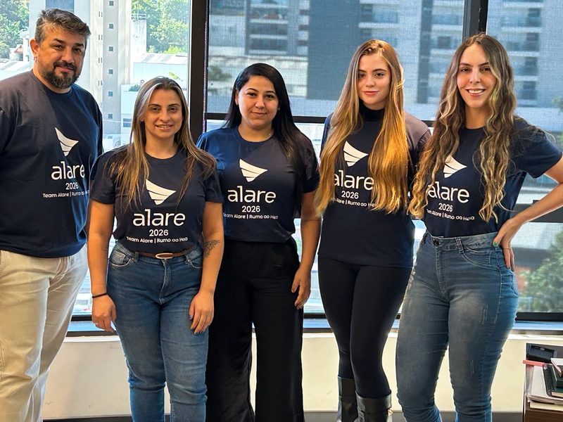 Cinco pessoas da equipe da Alare com a camiseta do time, em frente à janela do escritório
